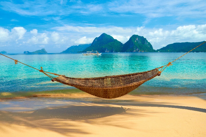 A hammock suspended over sand on a beach with clear blue water and lush green mountains in the background.