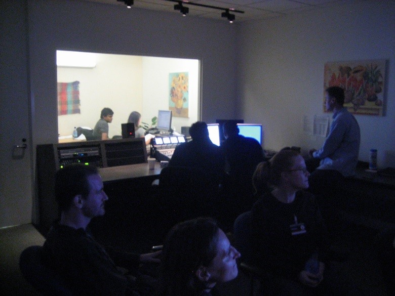 A photograph of a usability lab. Six people sit in a dark room with a large window leading to a brightly lit room with two others. Inside the dark room, three people are looking off-camera at a screen. Three others sit facing the window looking at a computer. The two people outside the window look at a computer.