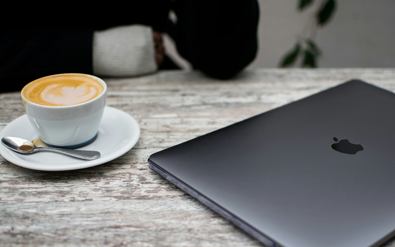 A photo taken from a slightly elevated angle shows a dark gray laptop and a small white teacup of coffee on a light-colored wooden table. A spoon rests on the saucer of the teacup, and a faint leaf or heart design is visible in the foam of the coffee. The top of the laptop is closed, with the apple logo visible.